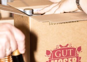 Zwei Hände packen Weinflaschen in einen offenen Karton mit der Aufschrift „(5+1 gratis) Weißburgunder Moserati Südsteiermark DAC 2023“. Eine Hand hält eine Flasche außerhalb des Kartons, was auf die Vorbereitung oder Verpackung für den Versand oder die Lagerung hindeutet.