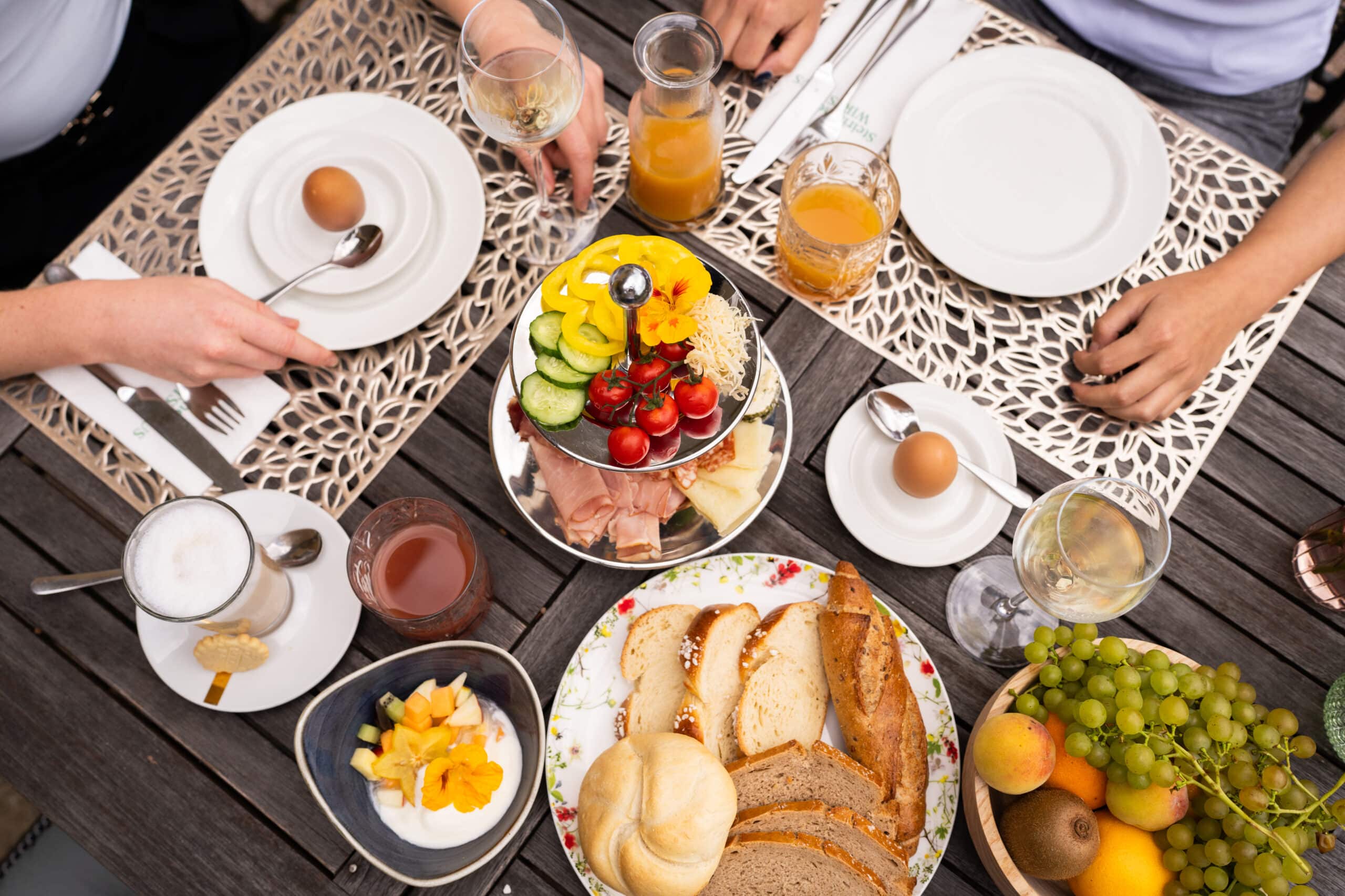 Zwei Personen sitzen an einem gedeckten Frühstückstisch im Winzerhotel am Gut Moser, an dem Brot, Eier, Obst, Gemüse, Saft und Kaffee auf gemusterten Tischsets stehen.