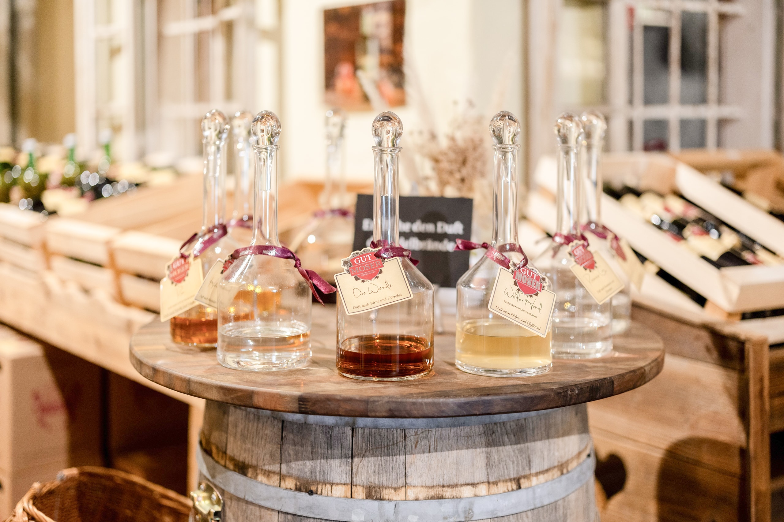 Mehrere Glaskaraffen, die mit verschiedenfarbigen Flüssigkeiten gefüllt sind, stehen auf einem rustikalen Holzfass, jede mit einem Etikett und einer Schleife versehen, in einem warm beleuchteten Laden mit Weinflaschen und Holzkisten im Hintergrund.