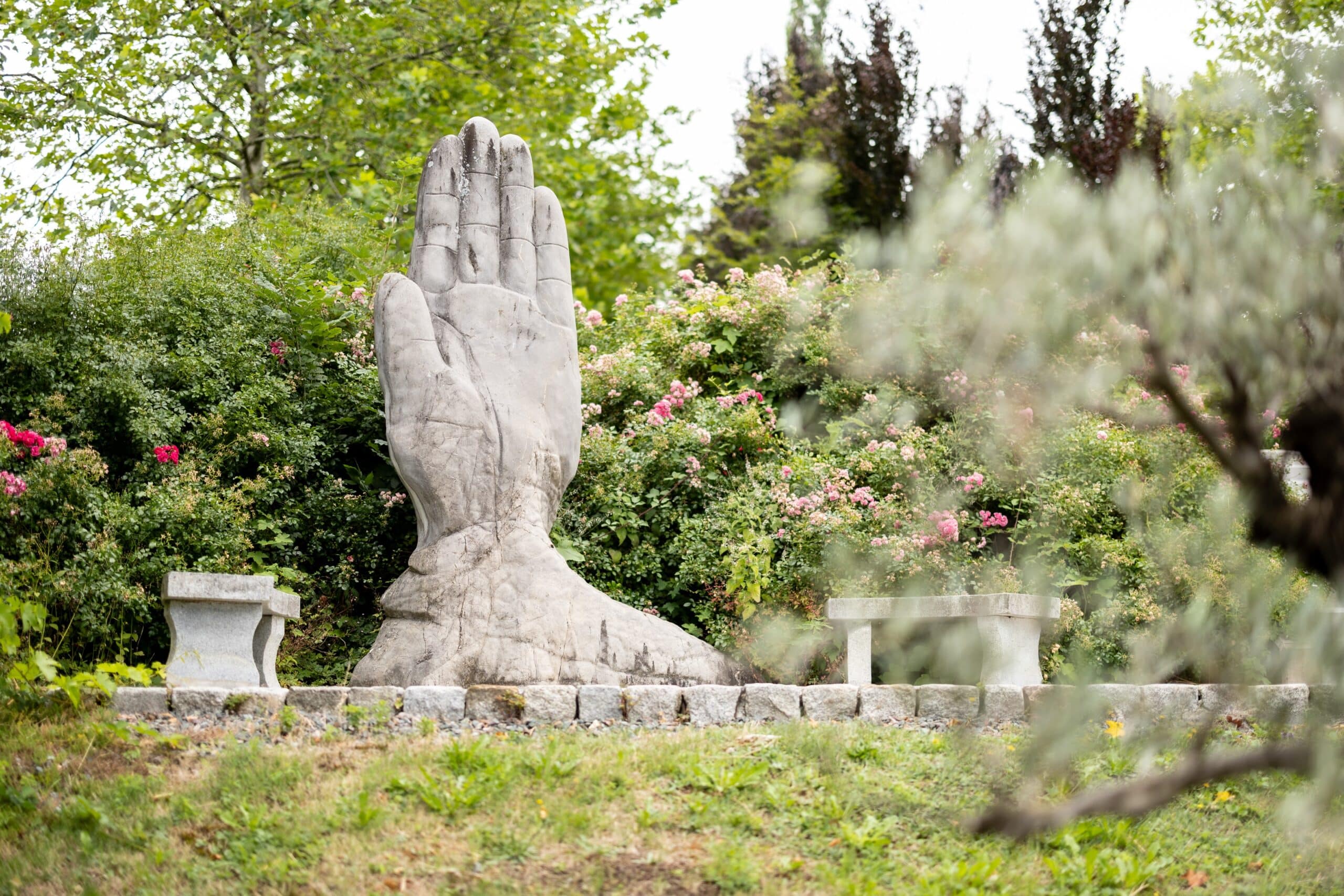 Eine große Steinskulptur einer aufrecht stehenden Hand ist von blühenden Sträuchern und Grün umgeben, daneben stehen zwei Steinbänke in einer Gartenanlage.