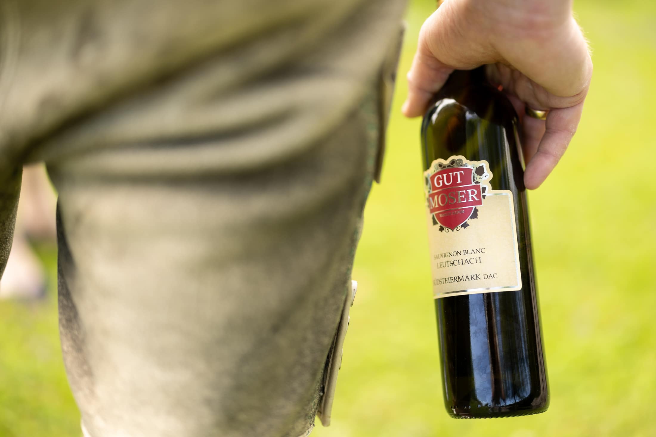 Eine Person in traditioneller Lederhose hält eine Flasche Wein mit der Aufschrift "Gut Moser" im Freien auf einer Wiese. Der Fokus liegt auf der Flasche und der Hand, mit unscharfem grünem Gras im Hintergrund.