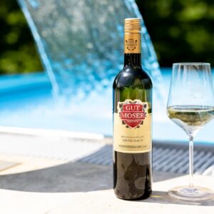 Eine Flasche Weißwein von Gut Moser und ein gefülltes Weinglas stehen am Rande eines Pools, mit einem Wasserspiel und blauem Wasser im Hintergrund, was auf eine entspannende Sommeratmosphäre hindeutet.