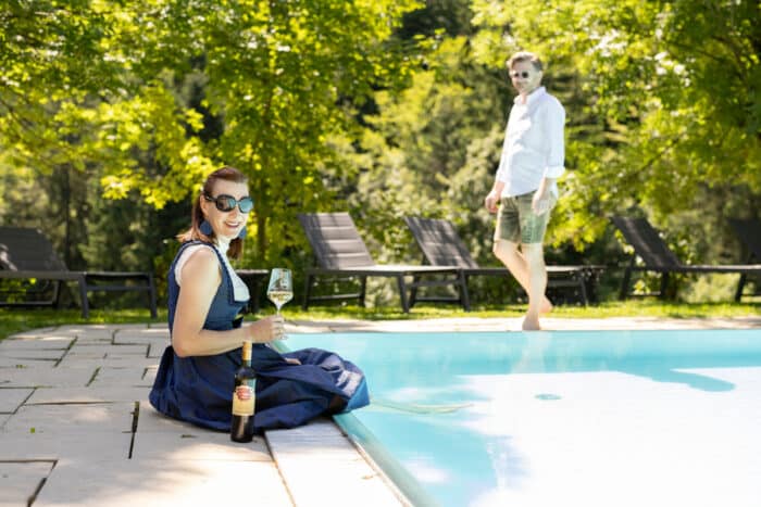 Eine Frau im Kleid sitzt am Pool, hält ein Glas Wein in der Hand und lächelt in die Kamera, daneben steht eine Flasche. Ein Mann in weißem Hemd und Shorts steht barfuß hinter ihr neben Liegestühlen und Grünpflanzen.