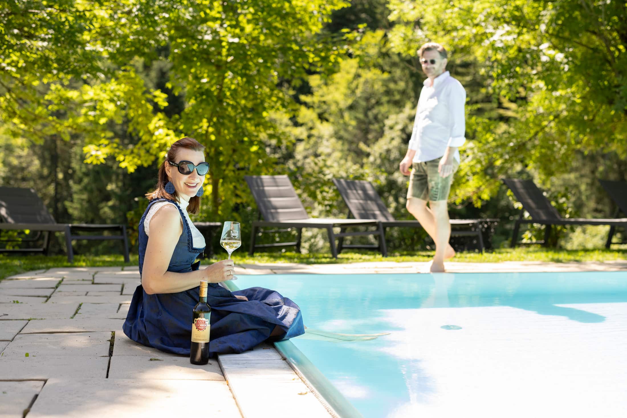 Eine Frau im Kleid sitzt am Pool, hält ein Glas Wein in der Hand und lächelt in die Kamera, daneben steht eine Flasche. Ein Mann in weißem Hemd und Shorts steht barfuß hinter ihr neben Liegestühlen und Grünpflanzen.