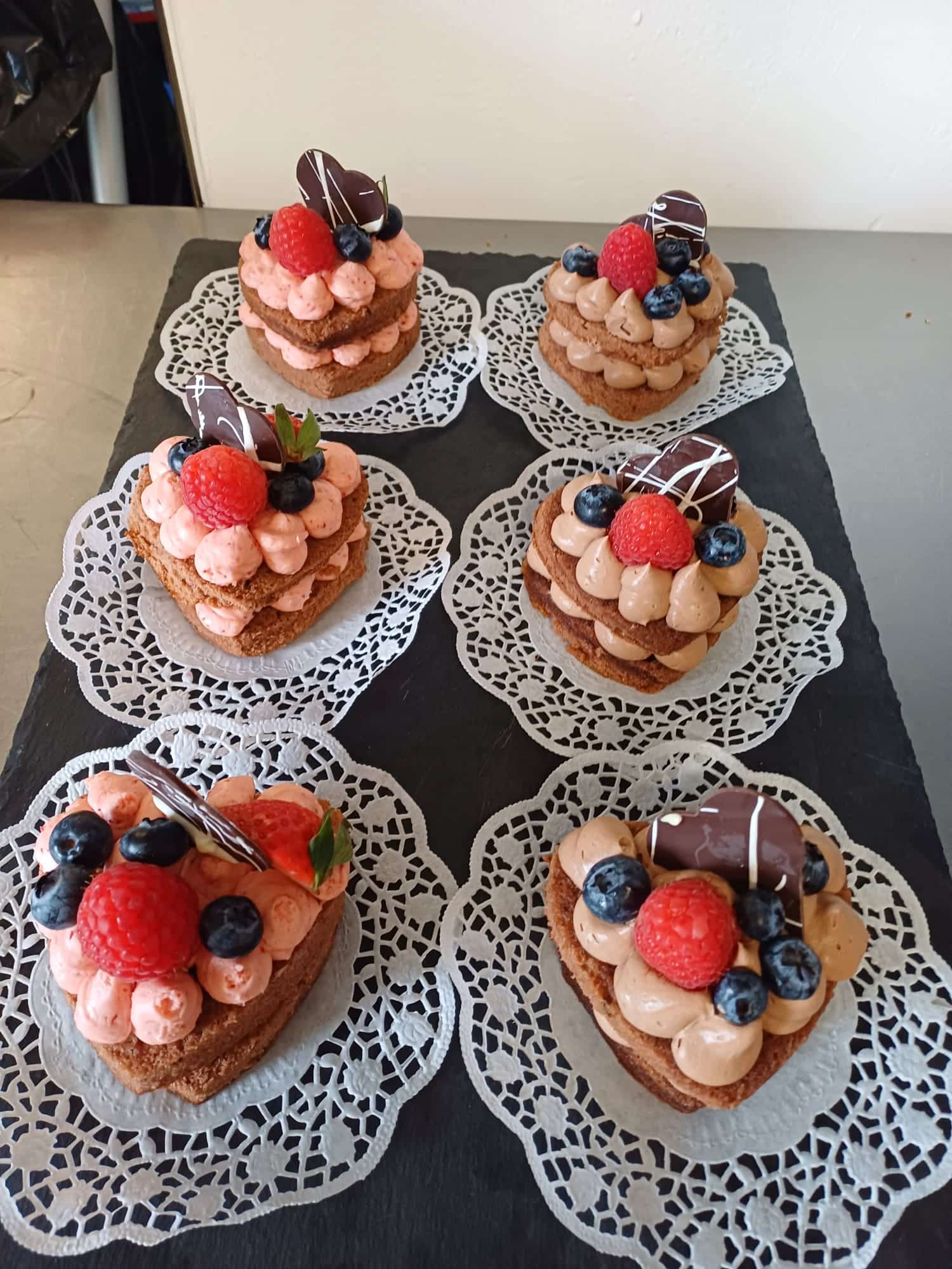 Sechs herzförmige Schokoladentörtchen mit rosa und brauner Sahne, Beeren und Schokolade, auf Spitzendeckchen auf einem Schiefertablett angerichtet.