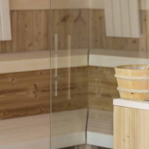 Holzsauna-Innenraum mit teilweise sichtbarer Glastür. Ein heller Holzeimer mit Schöpfkelle steht auf einer Bank. Die Saunawände sind mit vertikalen und horizontalen hellen Holzpaneelen verkleidet, die eine warme und einladende Atmosphäre schaffen.