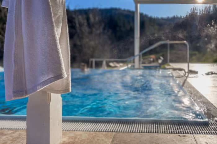 Ein weißes Handtuch hängt neben einem dampfenden Außenpool mit Bergen und Bäumen im unscharfen Hintergrund an einem sonnigen Tag. Warmwasser-Pool im Winzerhotel am Gut Moser.