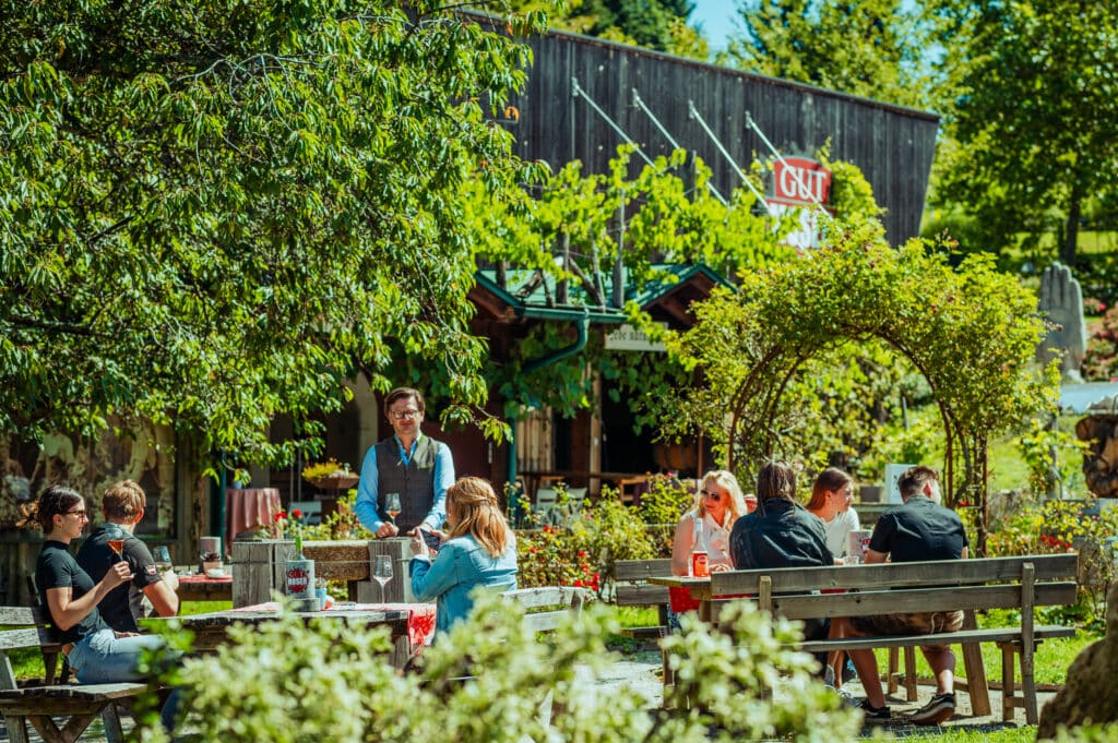 Menschen sitzen an Picknicktischen in einem üppigen Garten unter Bäumen, genießen Getränke und unterhalten sich im Restaurant. Eine Bedienung steht in der Nähe, während ein rustikales Holzgebäude im Hintergrund teilweise von Grün und einem "GUT"-Schild verdeckt ist.
