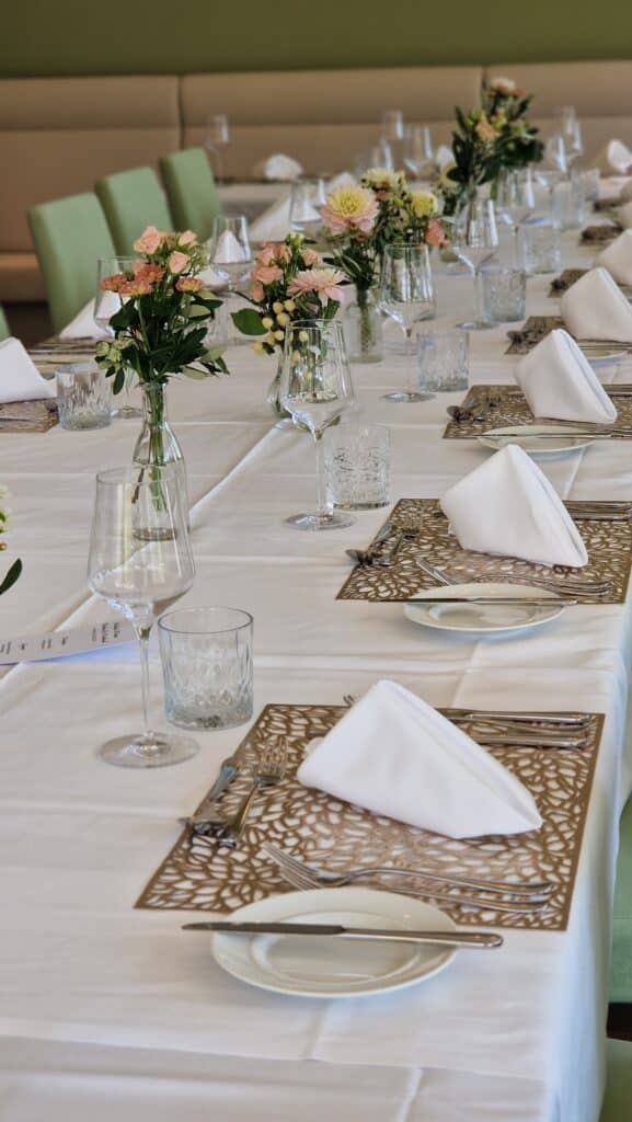 20250812_135416 Ein elegantes Restaurant-Tischset mit weißen Servietten, Gläsern, Blumengestecken und dekorativen Tischsets – bereit für ein festliches Essen.